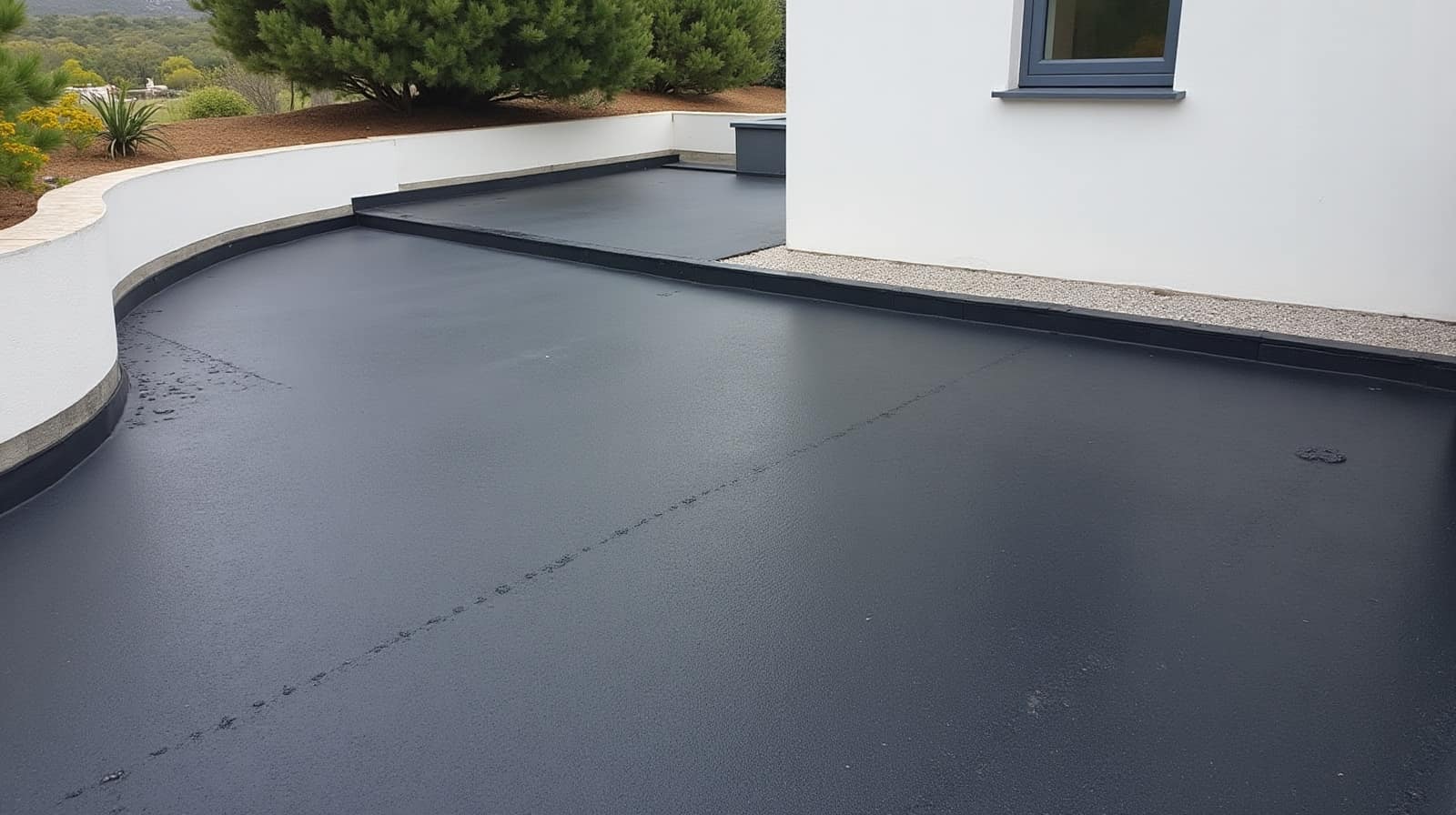 Toiture-terrasse à membrane sombre longée d'un acrotère blanc courbe, mur de maison et végétation attenants