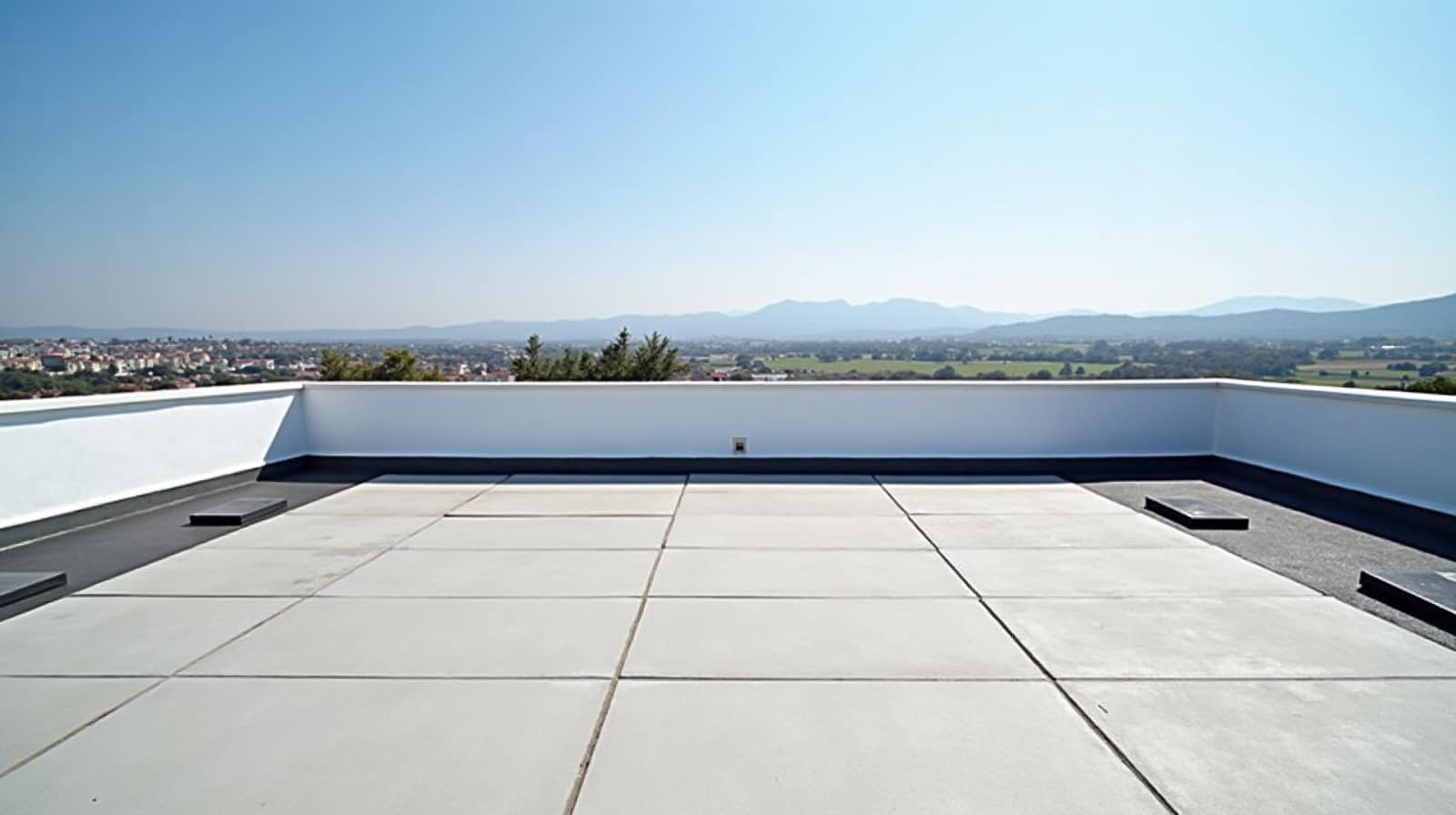 Terrasse de toit dallée en béton clair prolongée par la membrane sombre, acrotère blanc face à la plaine