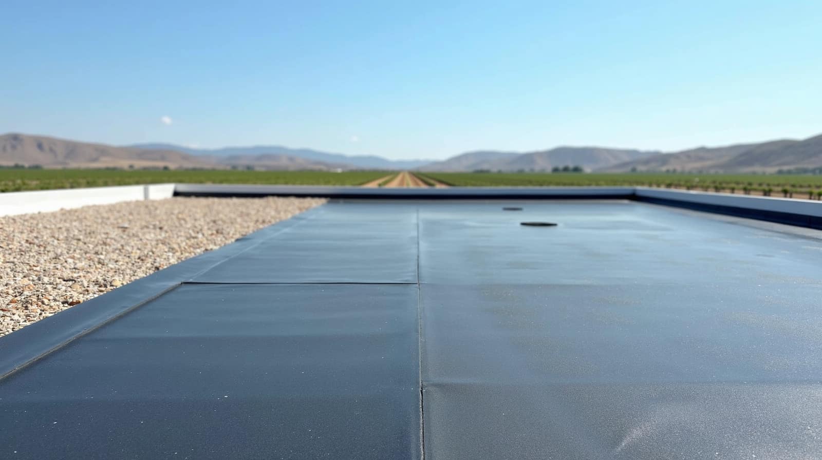 Toiture-terrasse à membrane sombre bordée de gravillon, ouverte sur la plaine cultivée et les collines
