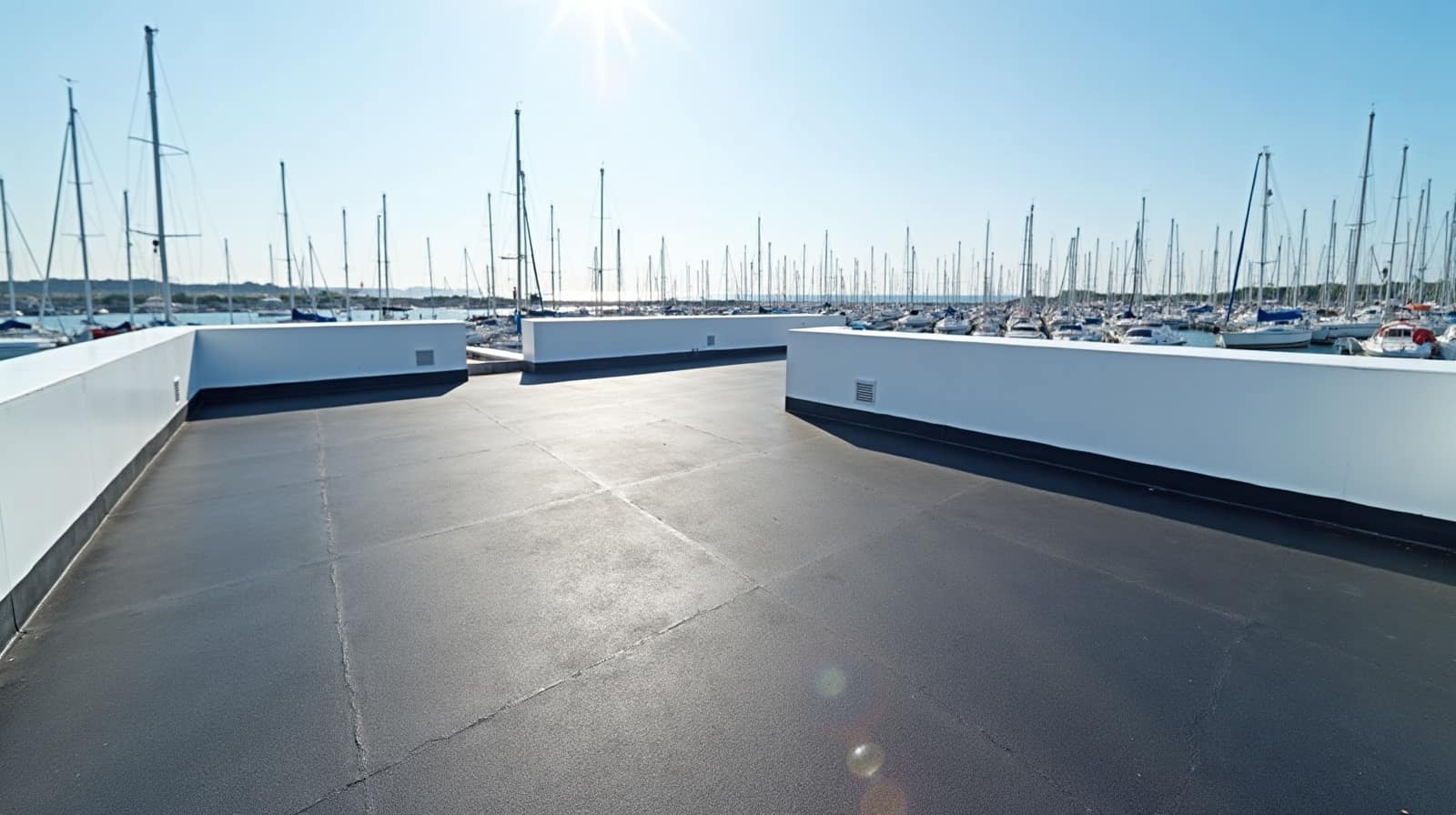 Toiture-terrasse sombre entourée d'acrotères blancs, mâts de voiliers du port au second plan